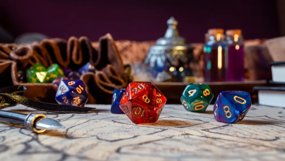 A red twenty-sided die beside blue and green dice on a map, with a pen and a leather dice bag